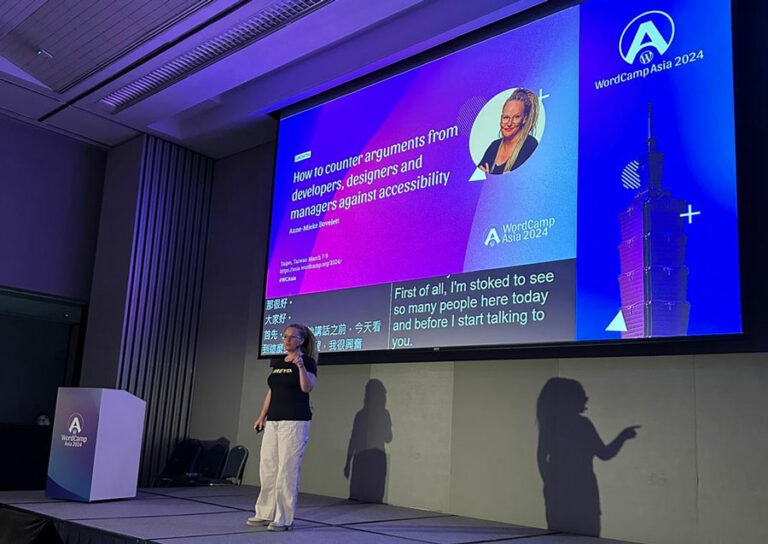 Anne on stage, wearing a huge blond ponytail, white trousers and a black shirt. On the screen behind her her talk called How to counter arguments against accessibility from desgners, developers and managers" is announced.