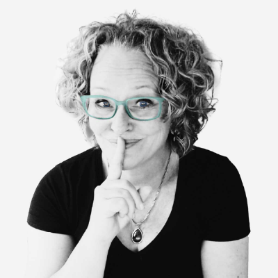 A portrait of Anne Bovelett. A woman with curly hair, turquoise glasses and a black top, smiling playfully while holding a finger to her lips to make a “shh” sound. The picture is black and white except for the glasses.